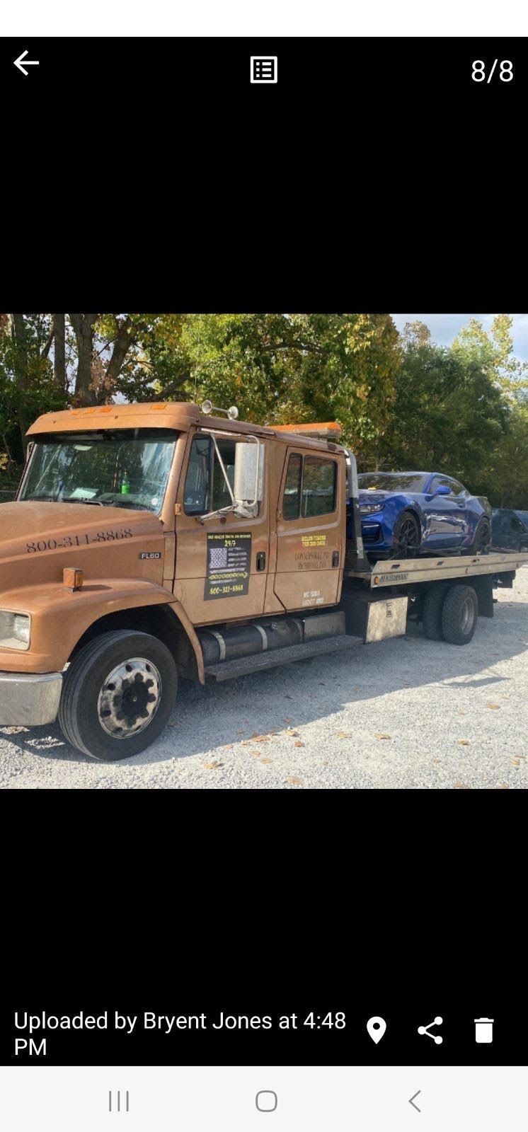 Tow truck carrying a blue sports car on a gravel road with trees in the background.