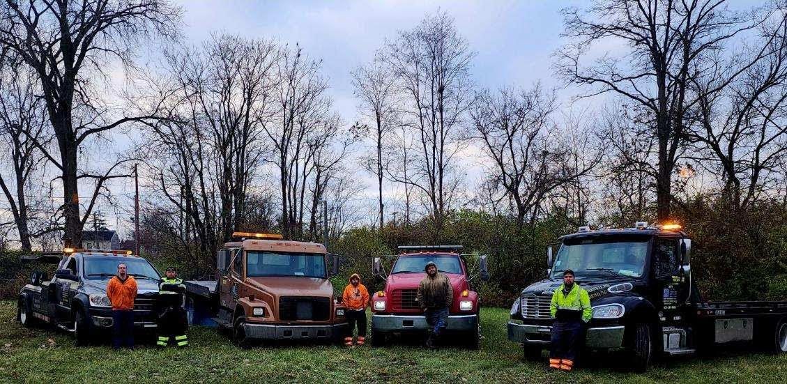 Four tow trucks lined up with drivers standing in front, surrounded by bare trees.