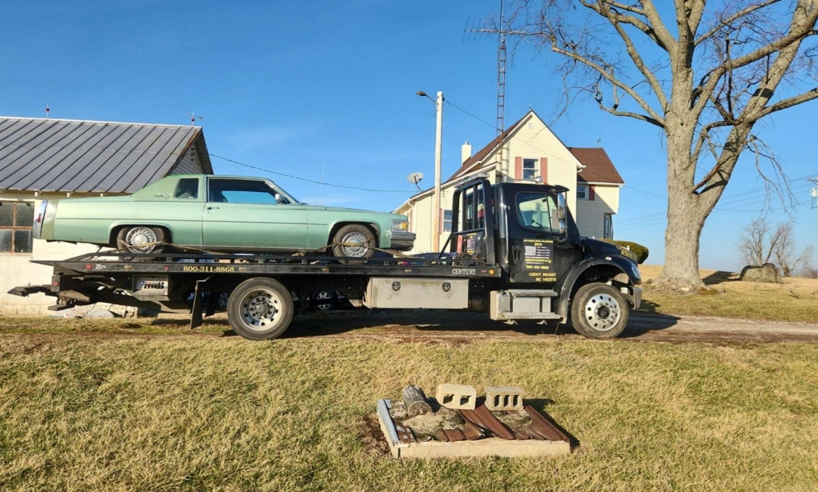 Tow truck transporting a vintage green car near a rural house and tree on a sunny day.