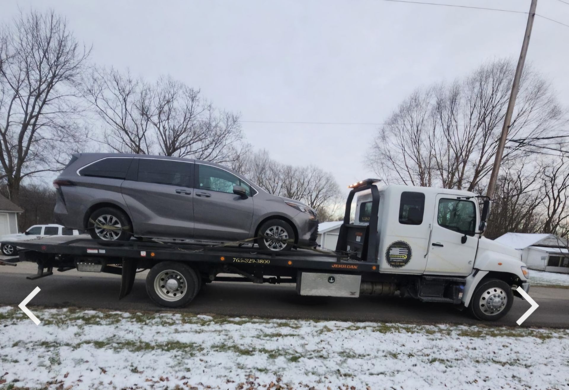 Tow truck transporting a gray minivan on a snowy residential street.