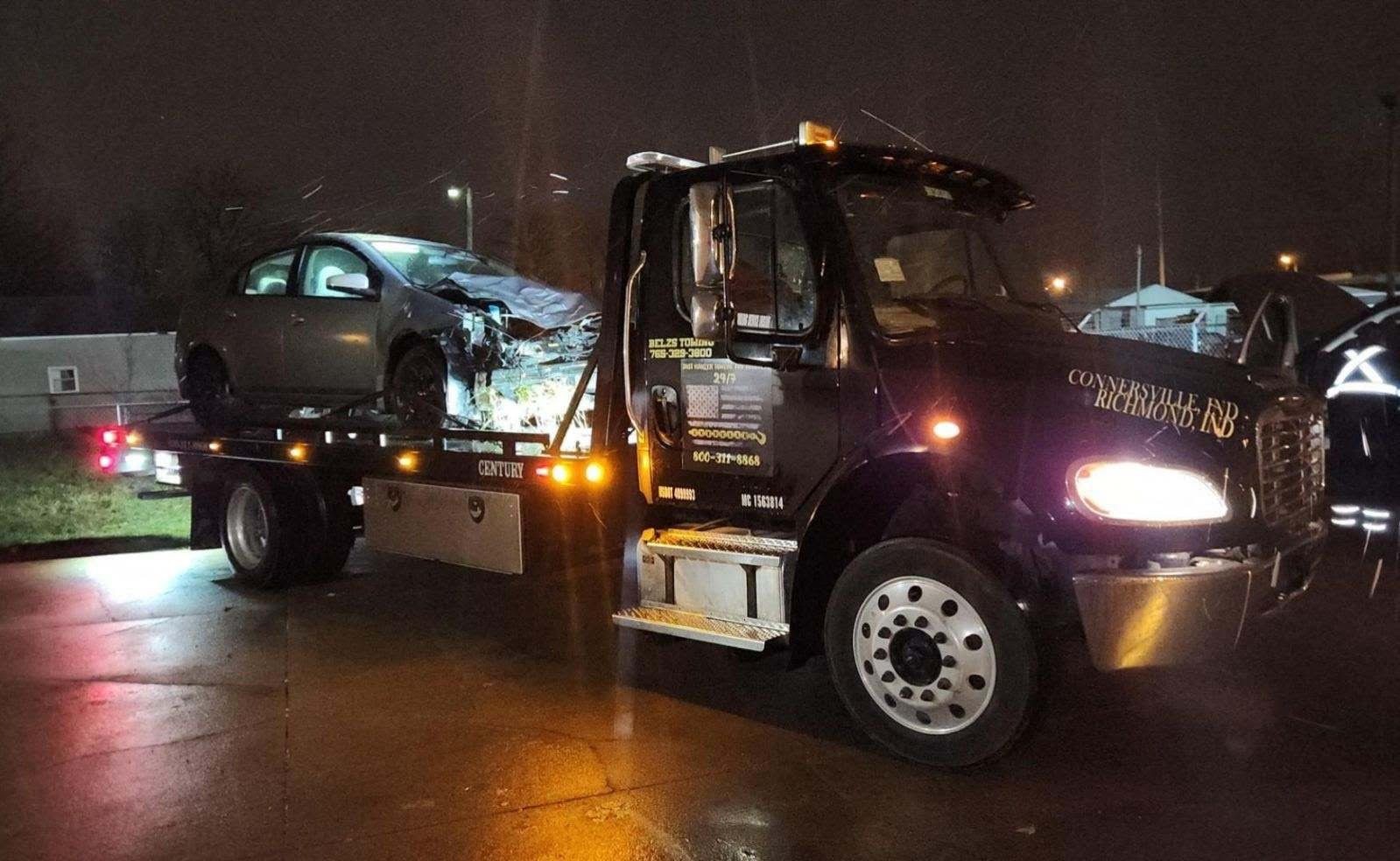 Tow truck carrying a damaged car at night with headlights on.