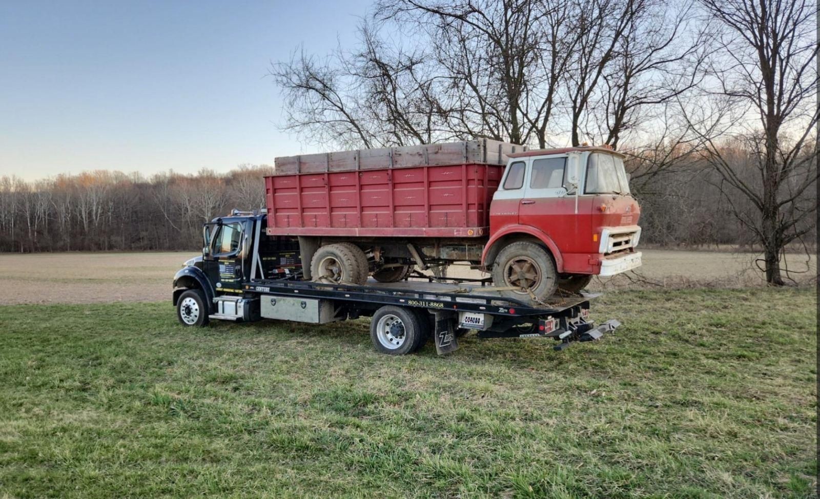 Tow truck carrying an old red farm truck on a flatbed in a grassy field.