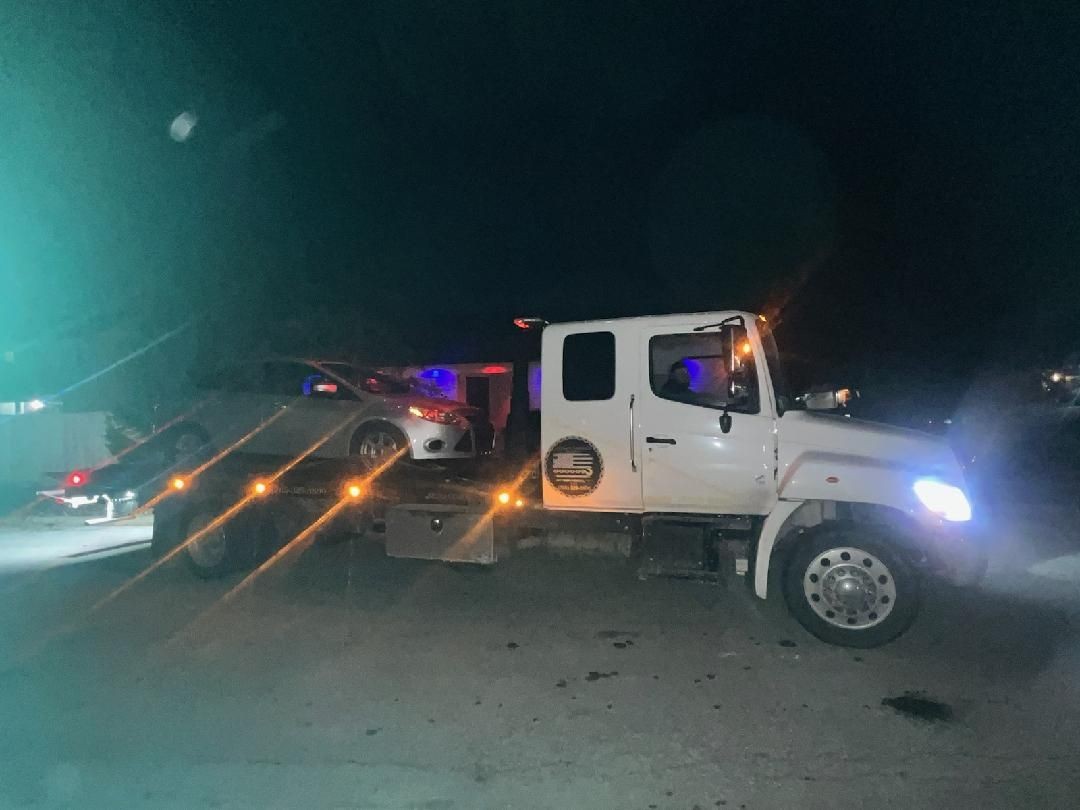 Tow truck transporting a white car at night, illuminated by the truck's lights.