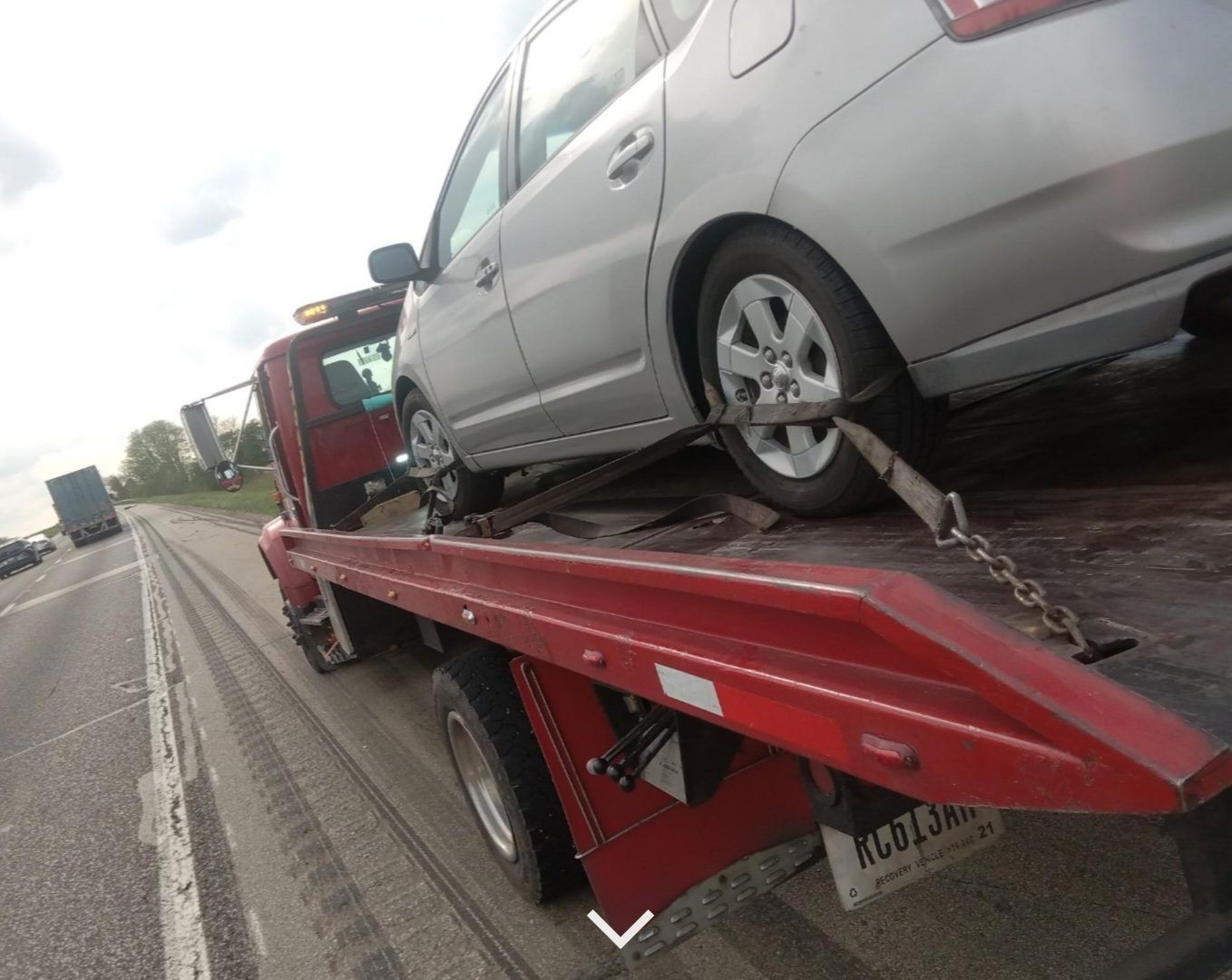 A silver car secured on a red tow truck driving on a highway.