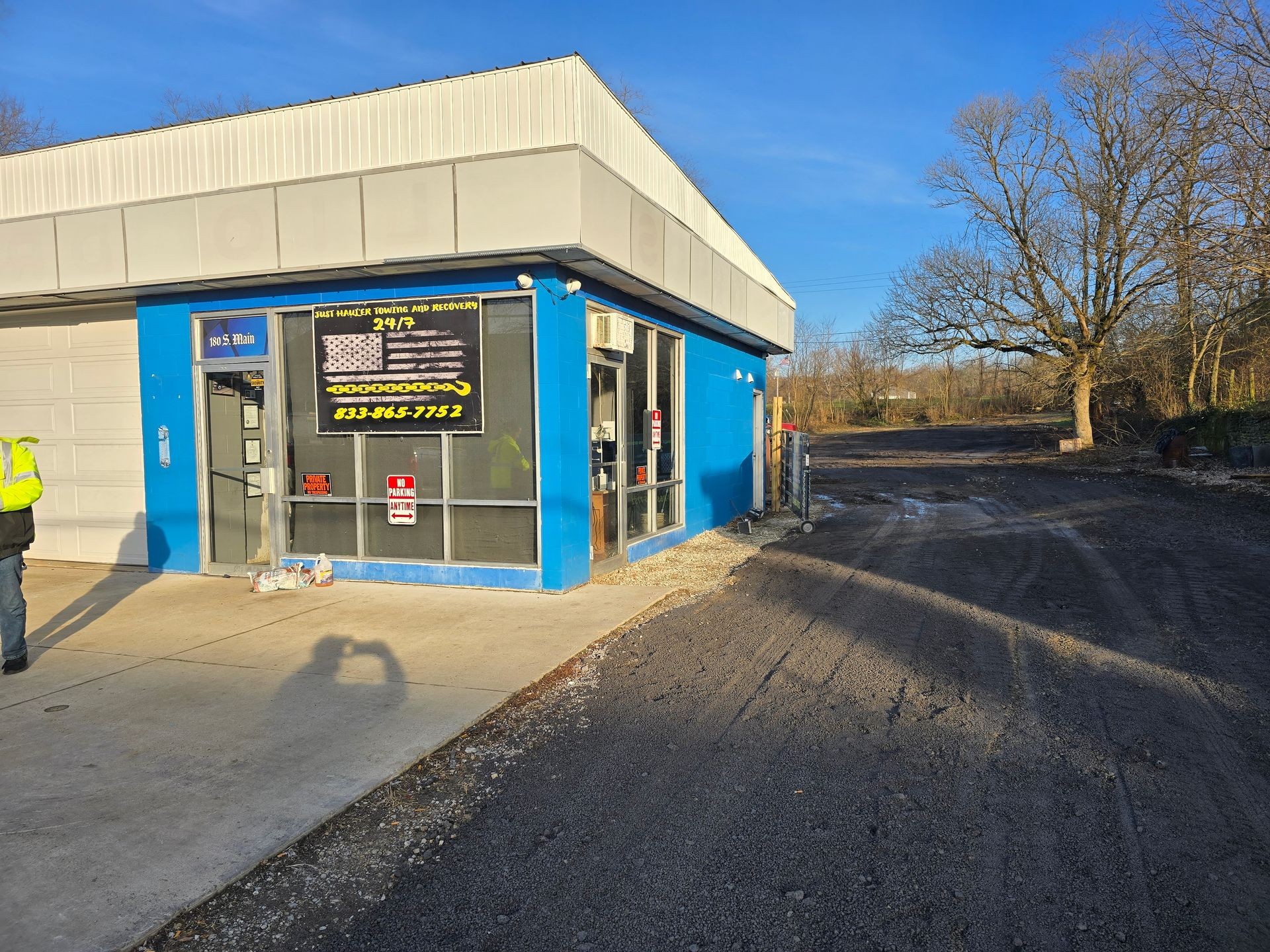 Exterior of a blue building with a towing company sign, surrounded by trees and a gravel driveway.