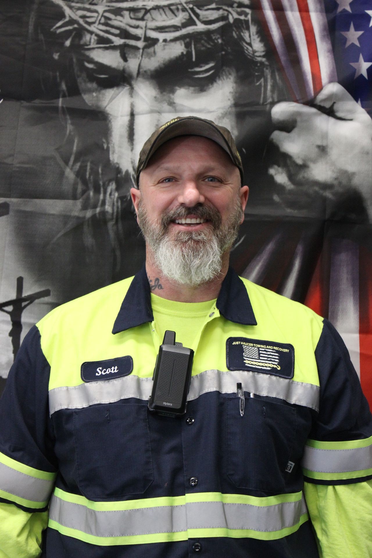 Man in reflective uniform with radio, smiling in front of American flag and religious-themed backdrop.