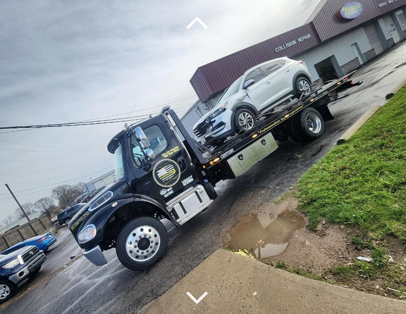 Tow truck carrying a silver SUV pulls up to a collision repair center on an overcast day.